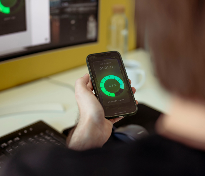 Auf dem Bild sieht man einen Mann von hinten sitzend an einem Schreibtisch vor einem Bildschirm. Er hat ein Smartphone in der Hand, auf dem die Driver Scoring App von prodot zu sehen ist.